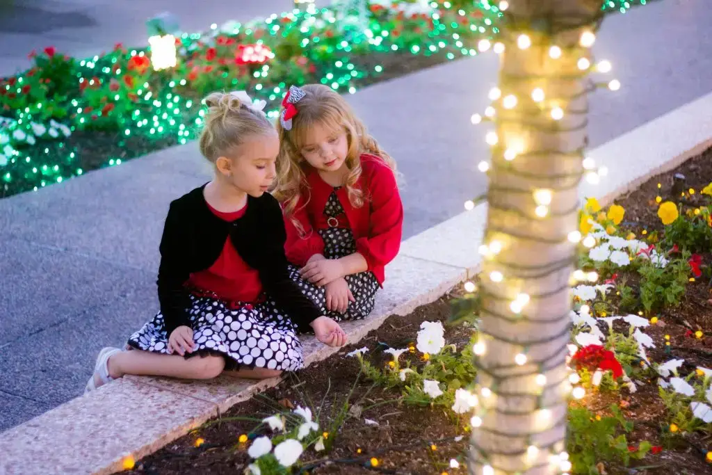 两个年轻女孩坐在梅萨神庙寺庙场地的花坛旁。床上可以看到圣诞彩灯,缠绕在棕榈树的树干上。